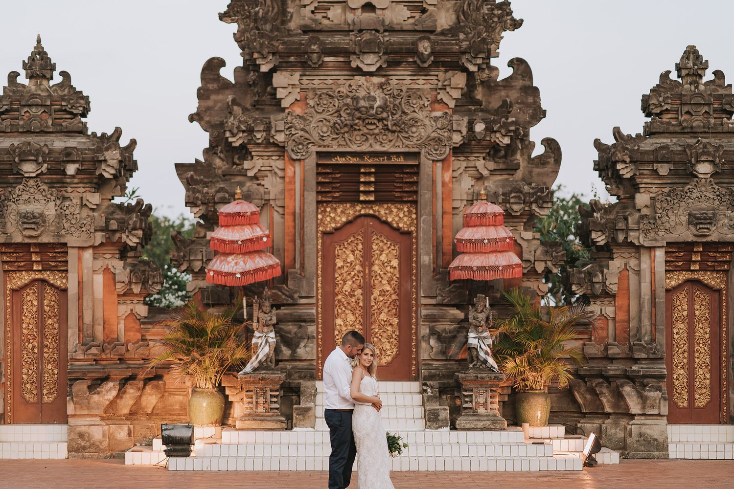 Couple embraces in front of ornate traditional Balinese temple gates with red umbrellas and intricate stone carvings.