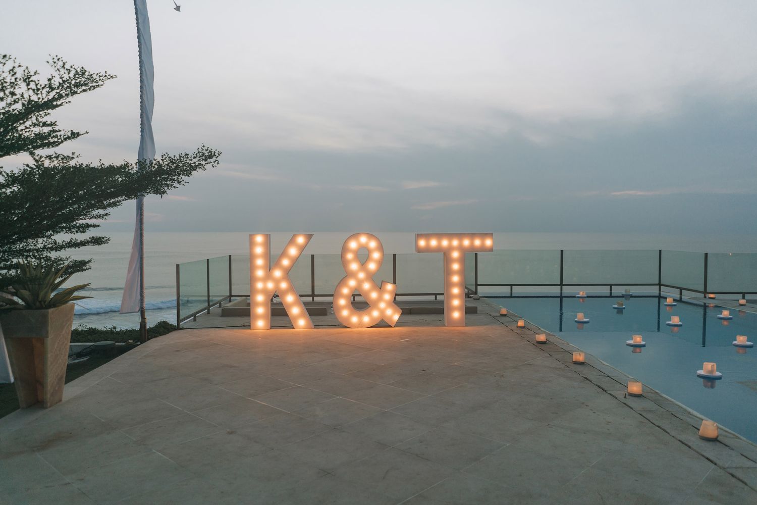 Illuminated marquee letters spell K&T on a seaside terrace with string lights and ocean views at dusk.