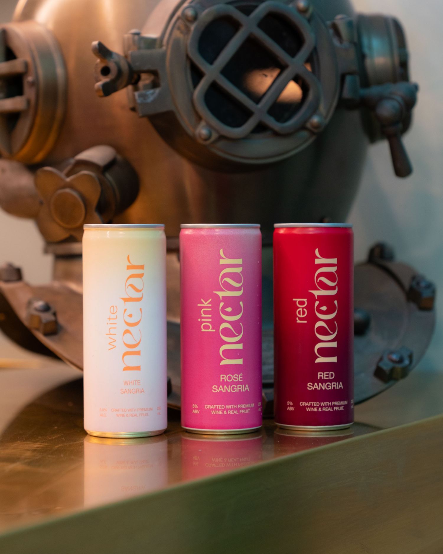 Three different flavors of Nectar canned drinks displayed on a metallic surface with industrial equipment behind.