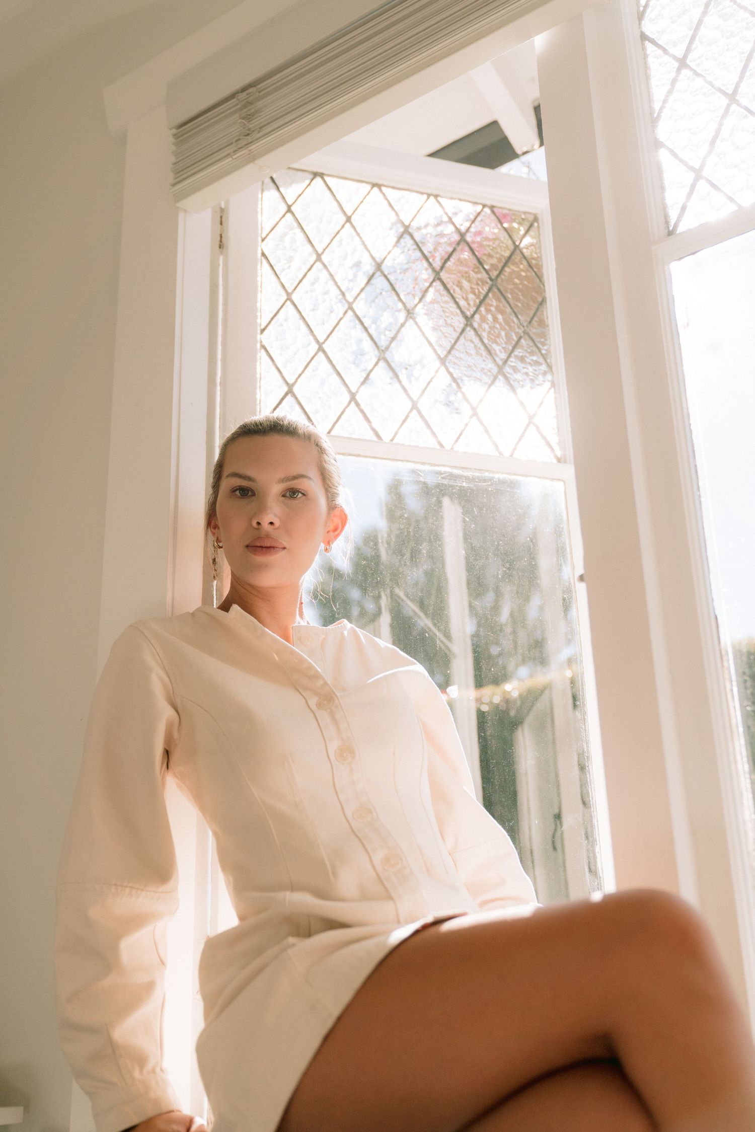 Woman in white linen shirt dress sitting on windowsill by leaded glass window in soft natural light.