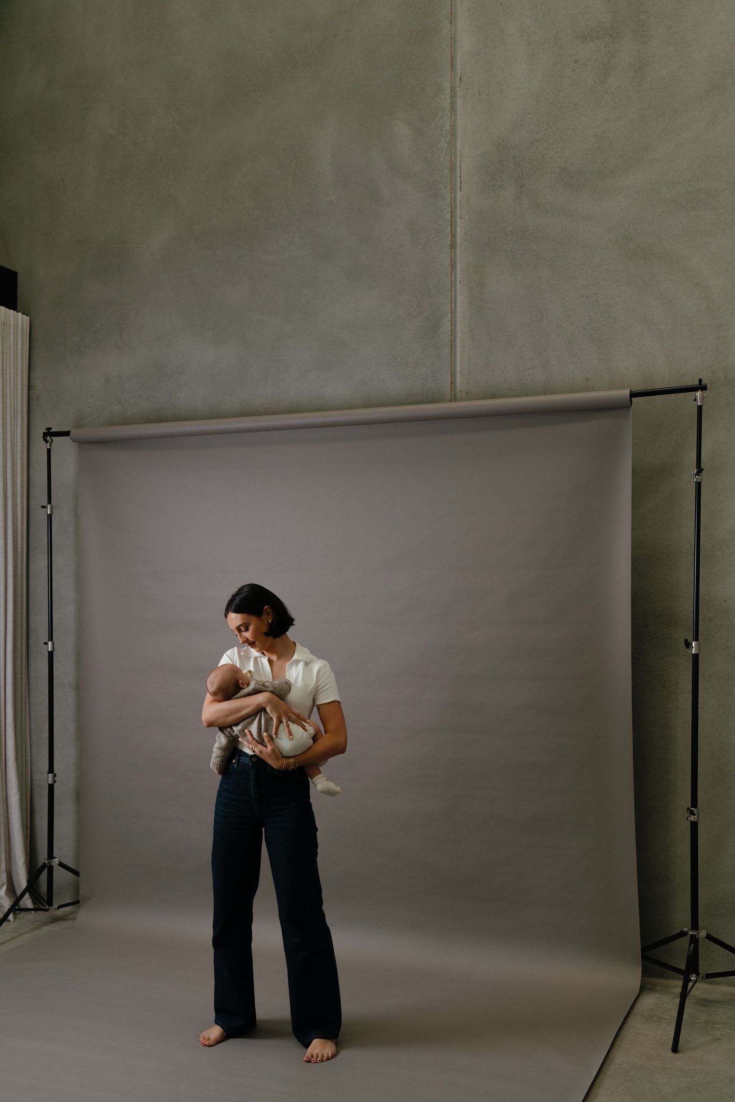 A woman stands barefoot in a photography studio holding a newborn baby, wearing a white shirt and dark wide-leg pants.
