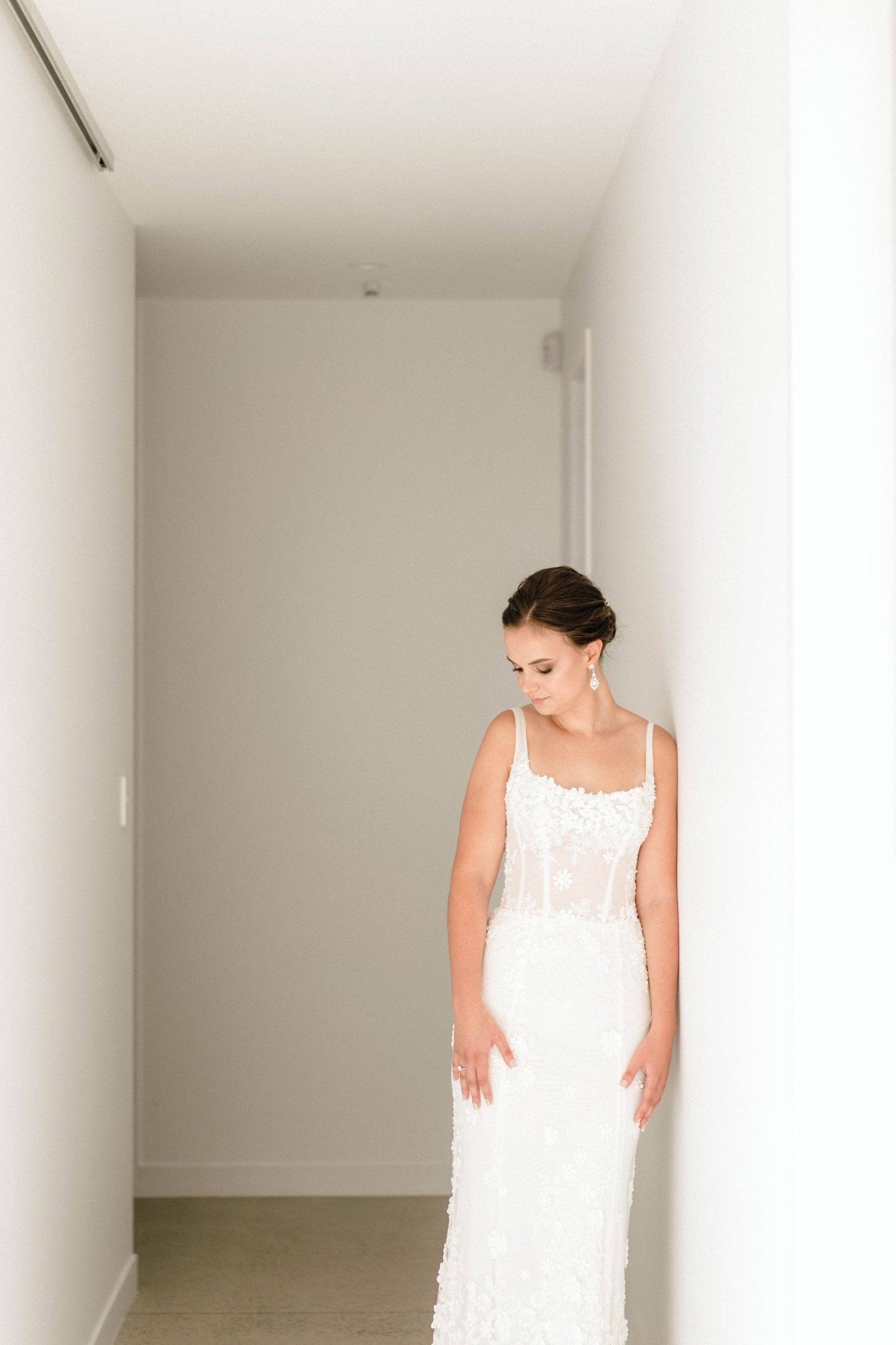 A woman in an elegant white lace wedding dress stands thoughtfully in a minimalist white hallway.
