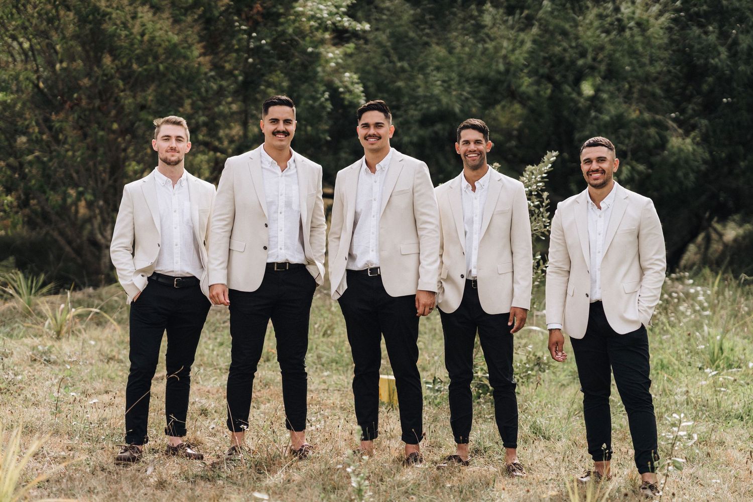 Groomsmen in black and white formal attire pose together against a natural backdrop.