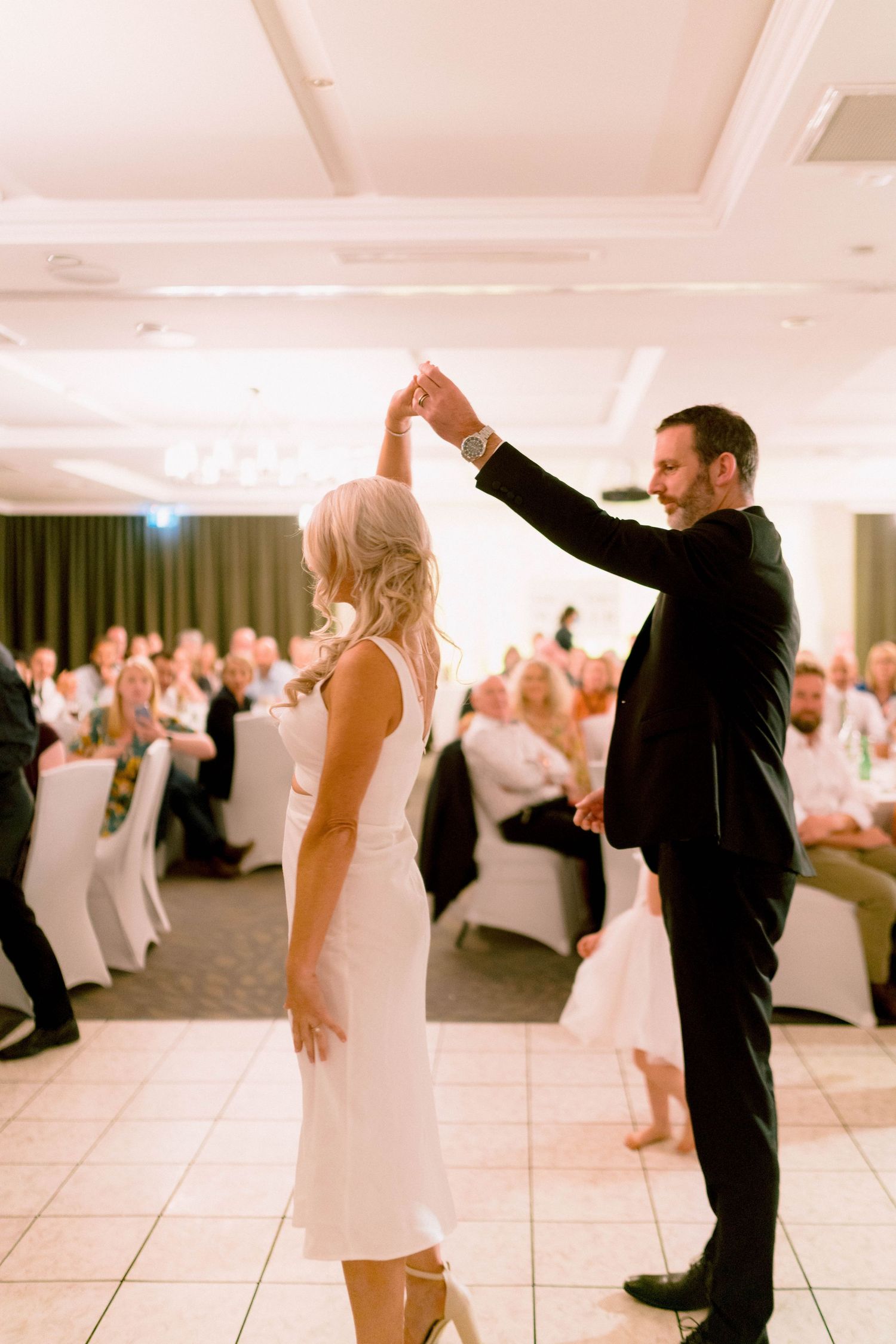 A couple shares their first dance on the ballroom floor surrounded by wedding guests in white chairs.