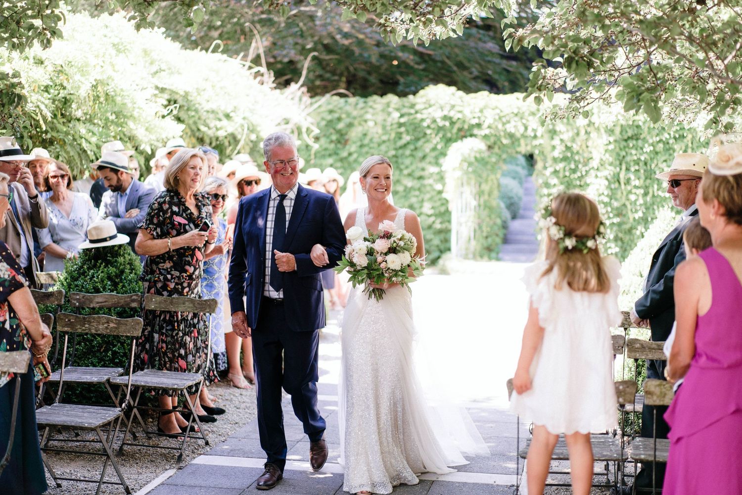 Wedding ceremony processional under a lush garden archway with guests seated along the aisle.