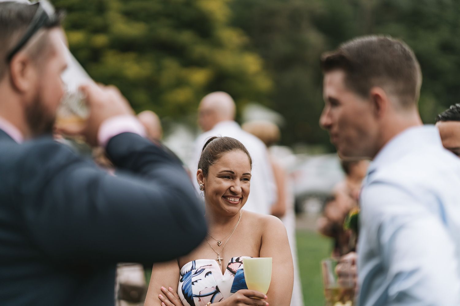 A person holding a small wrapped gift box smiles while guests mingle at an outdoor reception.
