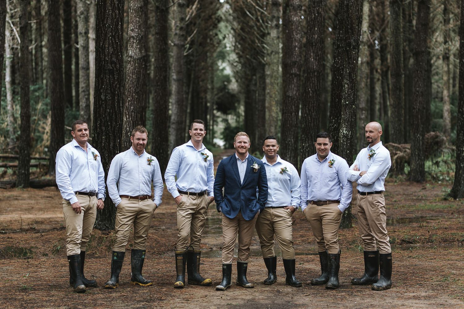 Wedding party wearing matching light blue shirts and khaki pants pose together in a pine forest setting.