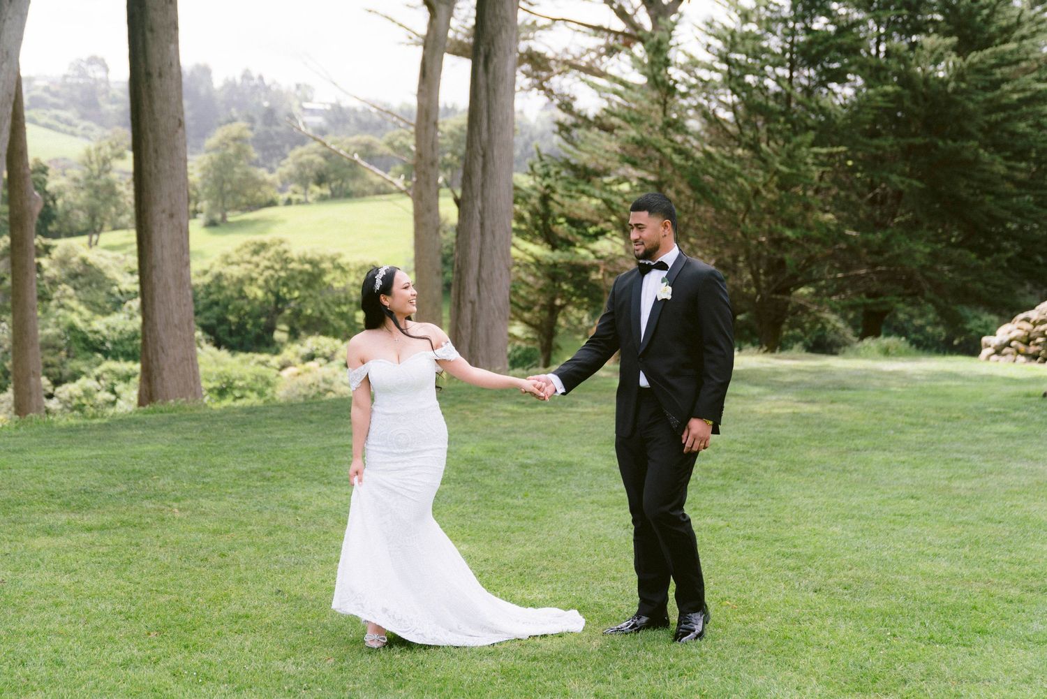 Wedding couple poses for romantic photos on a grassy hillside surrounded by evergreen trees.