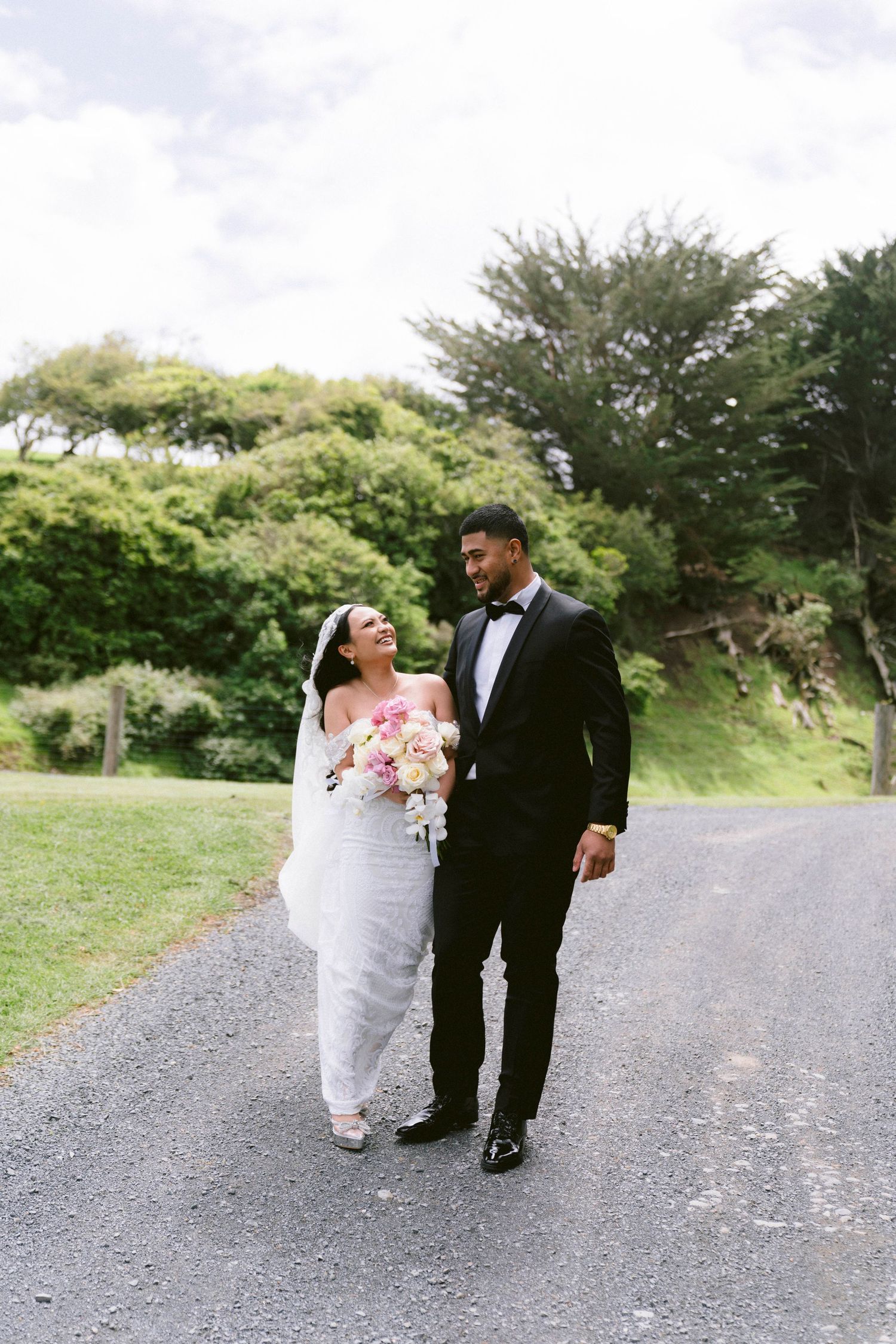 A couple walks down a gravel path on their wedding day, surrounded by lush greenery.