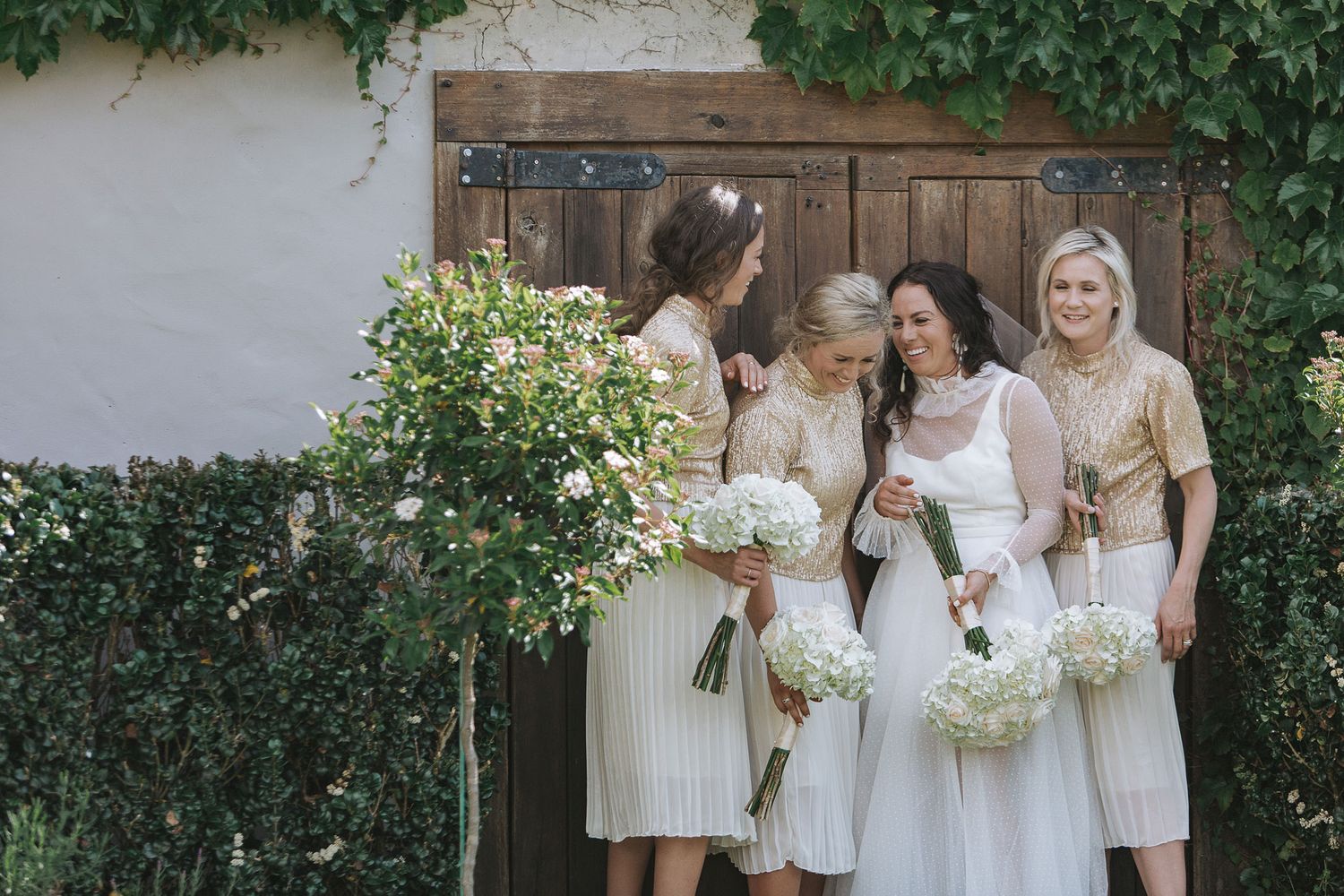 A bridal party in cream-colored dresses stands against a rustic wooden door with greenery and floral bouquets.