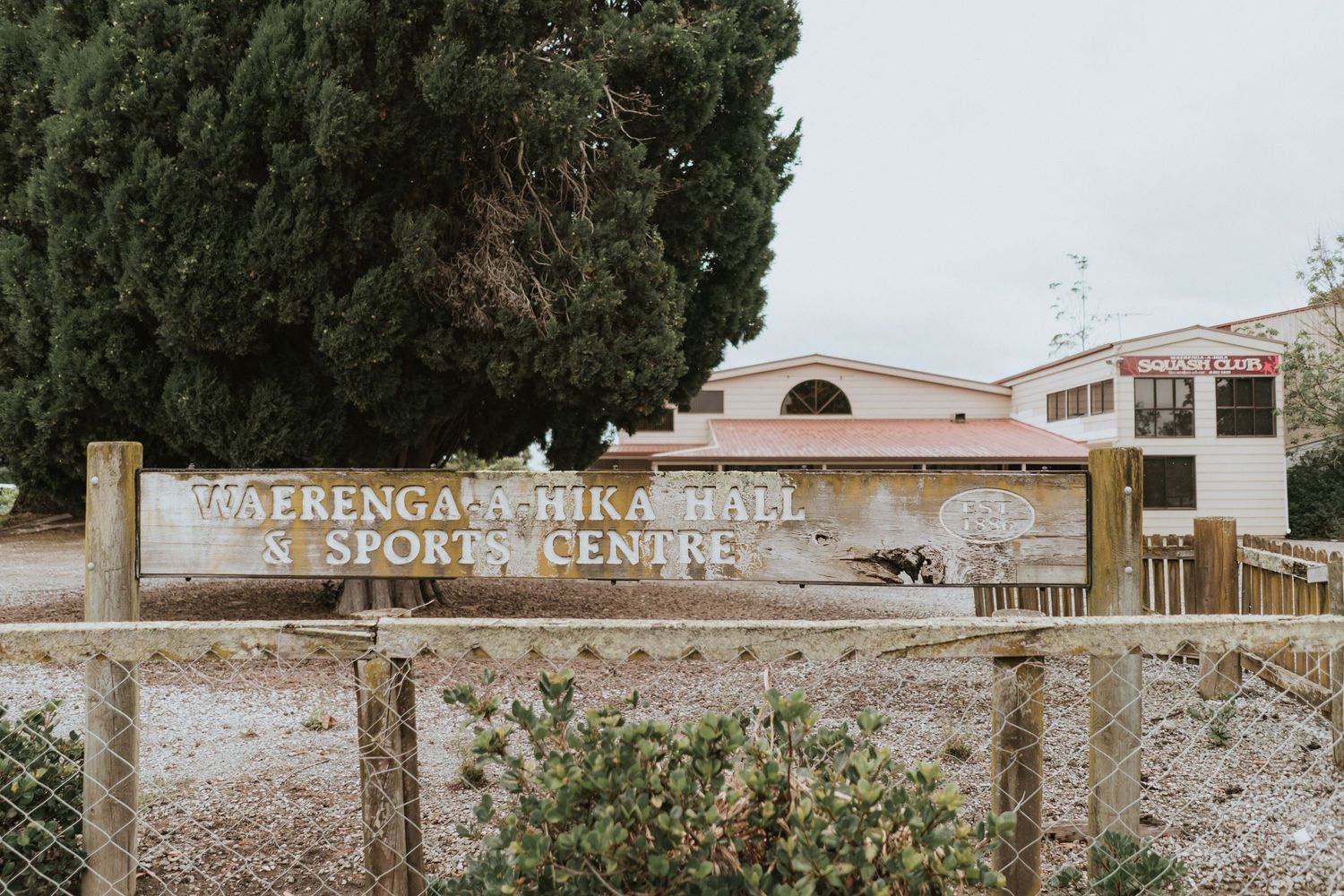 A weathered sign for a Spanish mission building with stone walls and arches stands behind metal fencing.