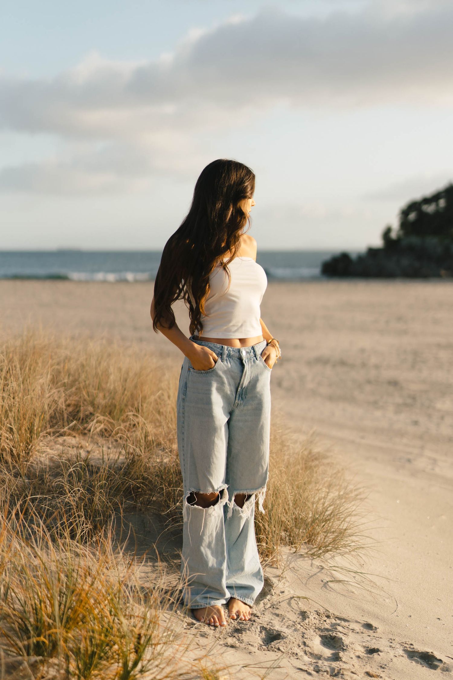 Figure in white top and ripped jeans poses on beach with ocean view.