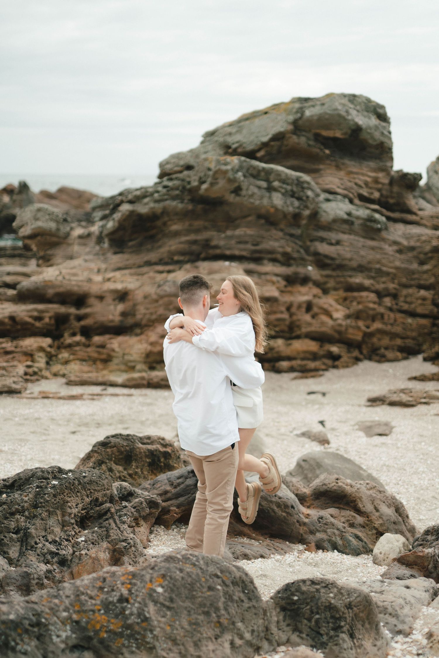 Couple in white embracing on a rocky beach landscape.