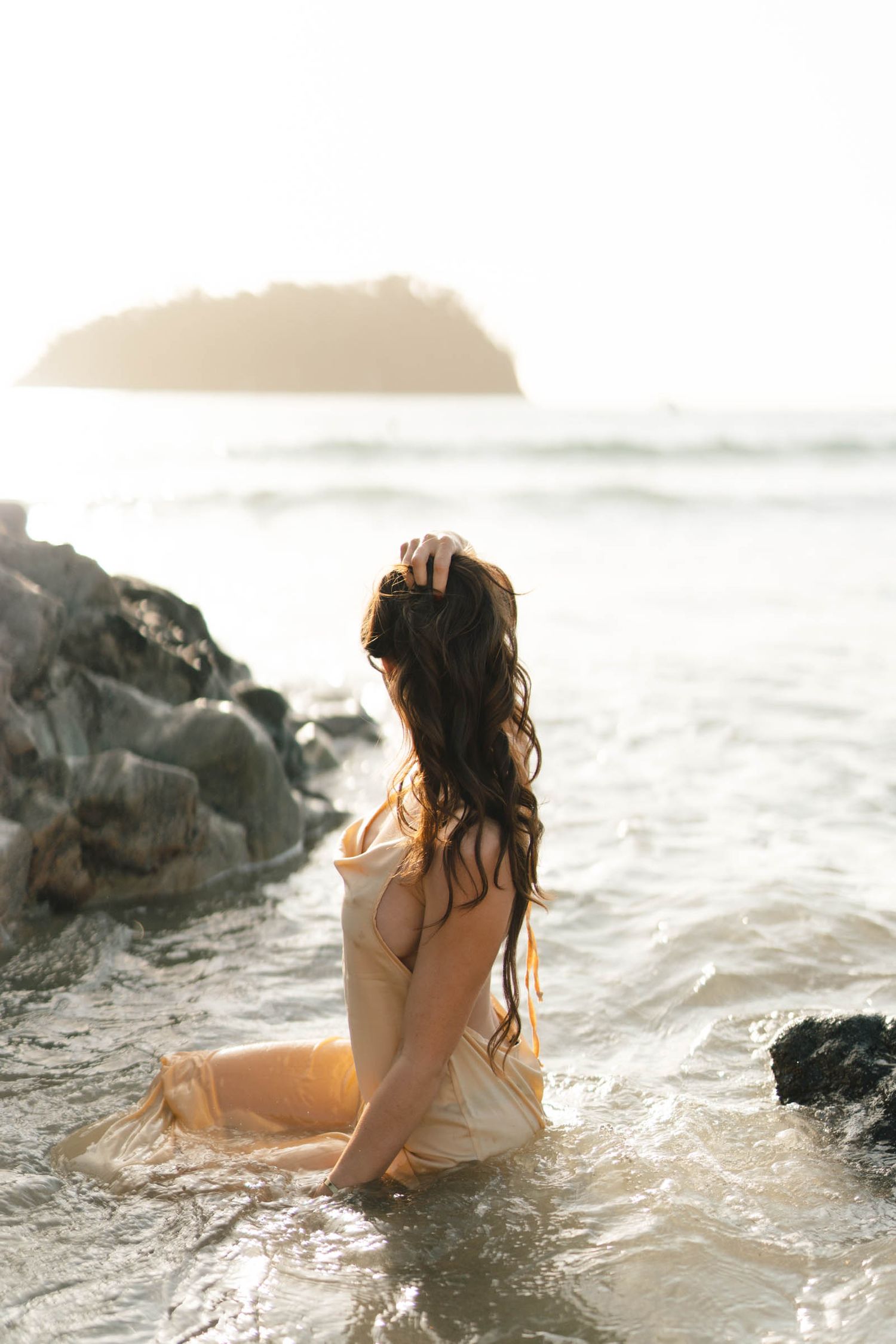 A person in a light dress sits among rocks in the ocean during sunset.