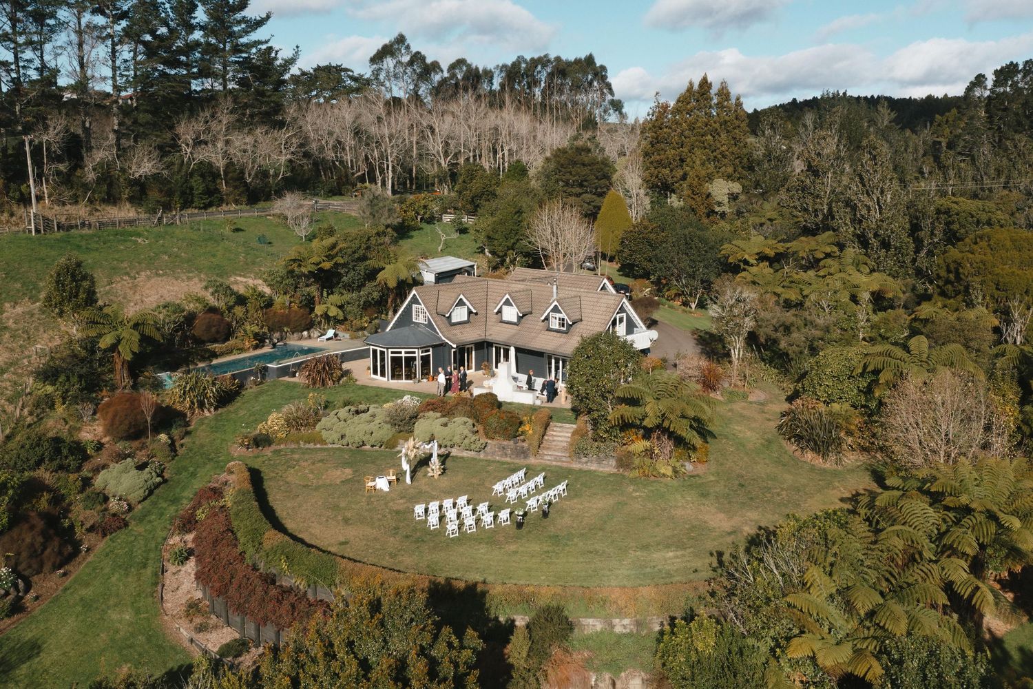 Aerial view of a white cottage with wrap-around porch nestled on a hillside surrounded by lush gardens and white chairs.