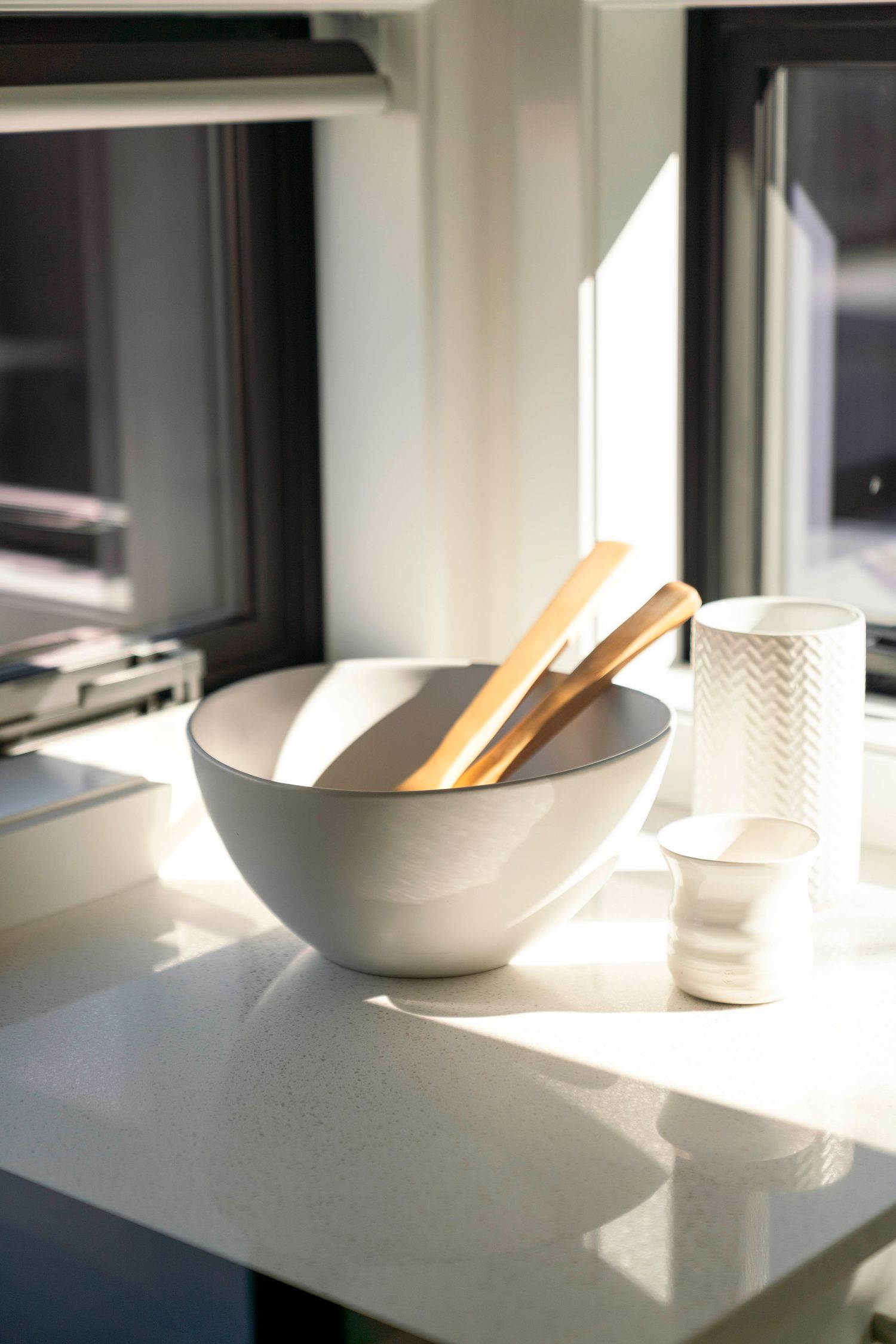 White ceramic bowl with wooden utensils sits on a bright sunlit counter next to a textured glass.