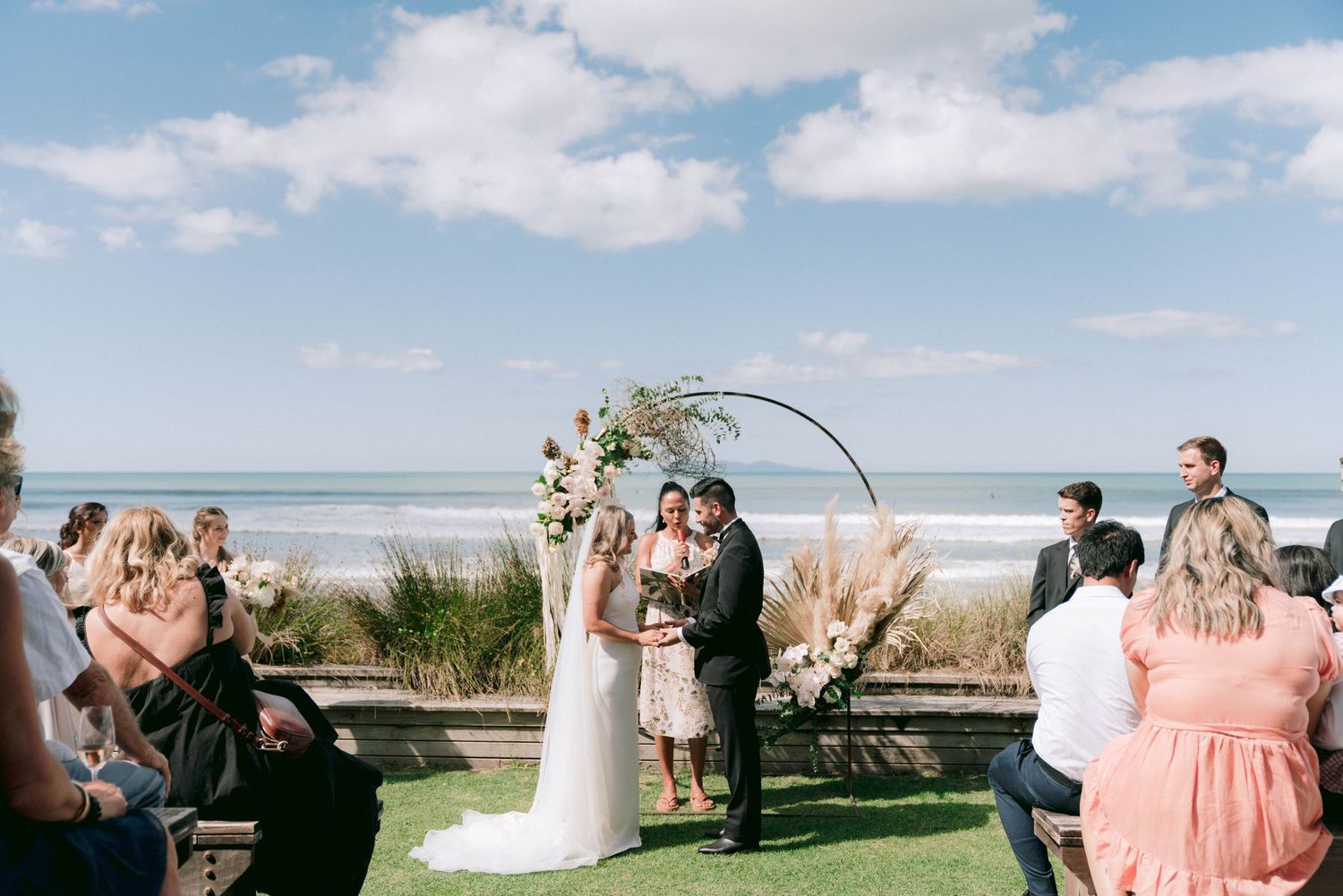 Wedding party gathering on a beach with bridesmaids in pink dresses under bright blue skies.