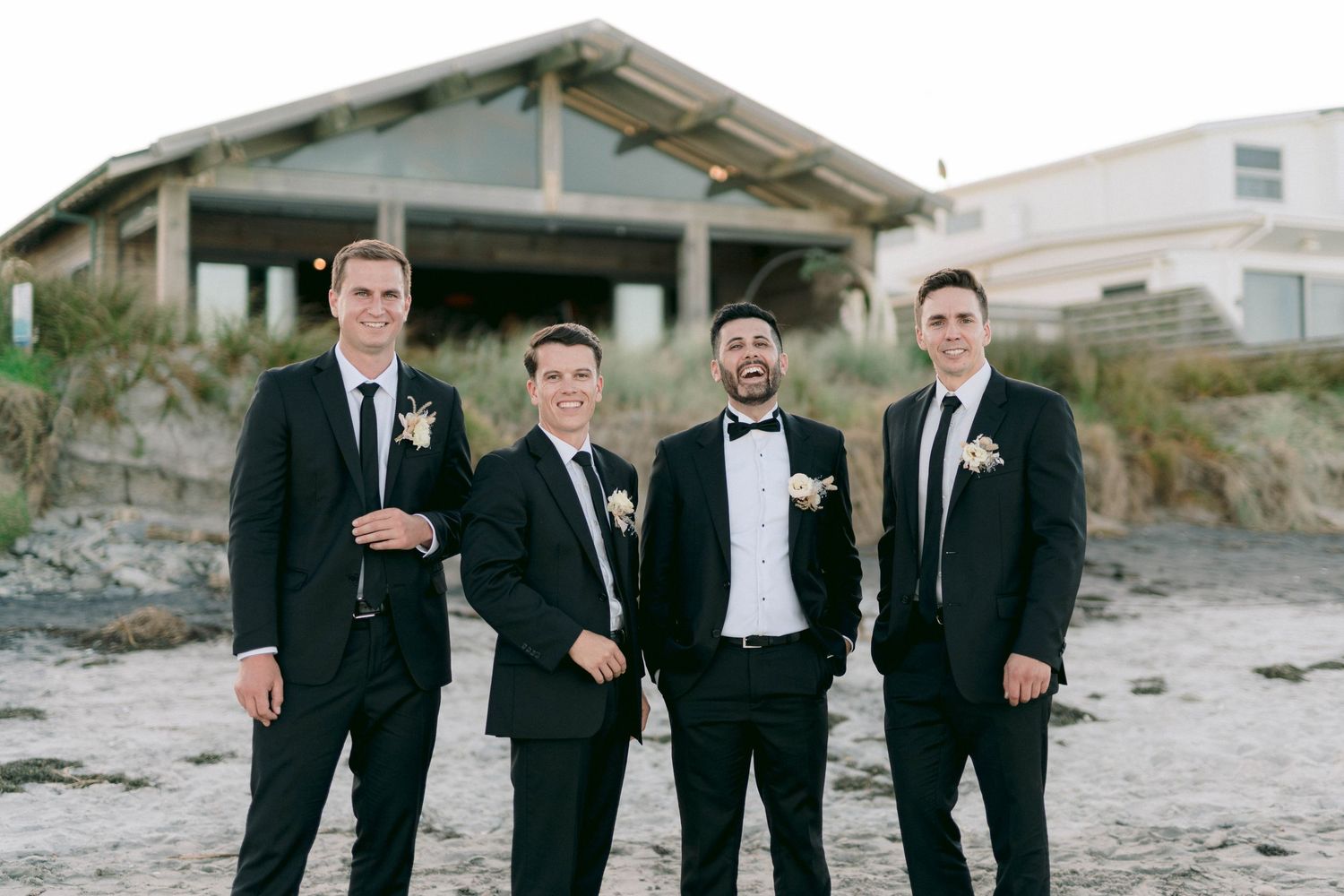 Four groomsmen in dark suits pose together on a beach at sunset.