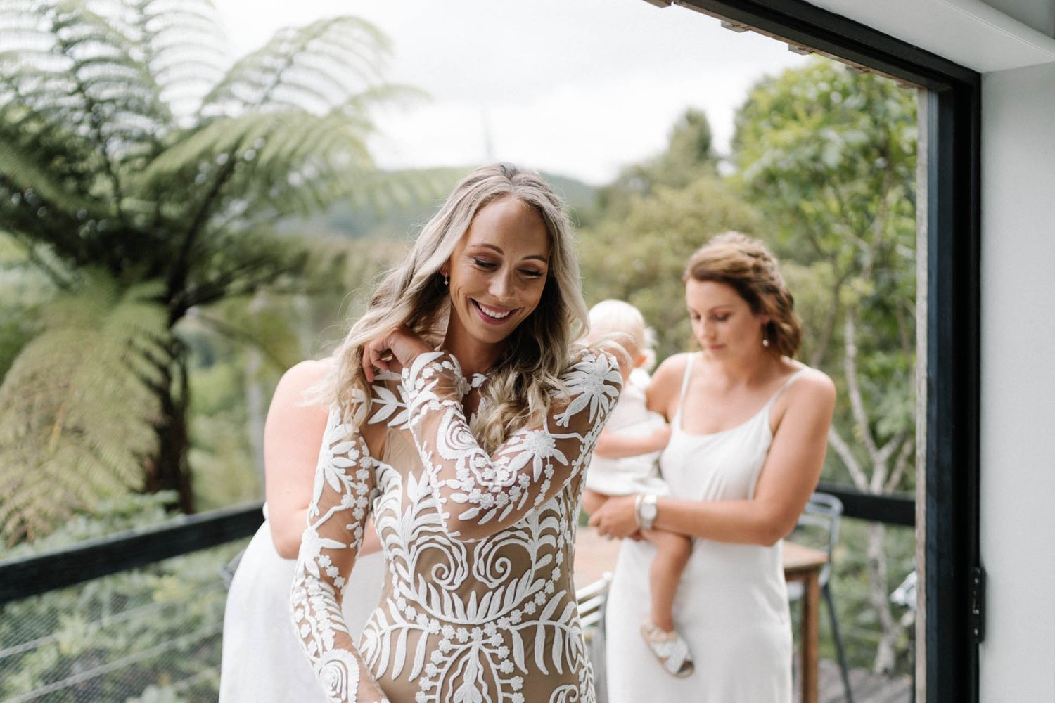 Elegant white lace wedding dress with intricate detail being fitted in a bright indoor space with ferns.