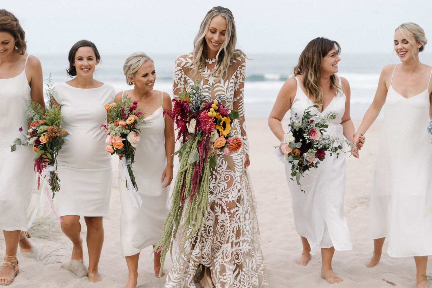 A beachside wedding party walks together in white dresses carrying colorful bouquets along the shoreline.