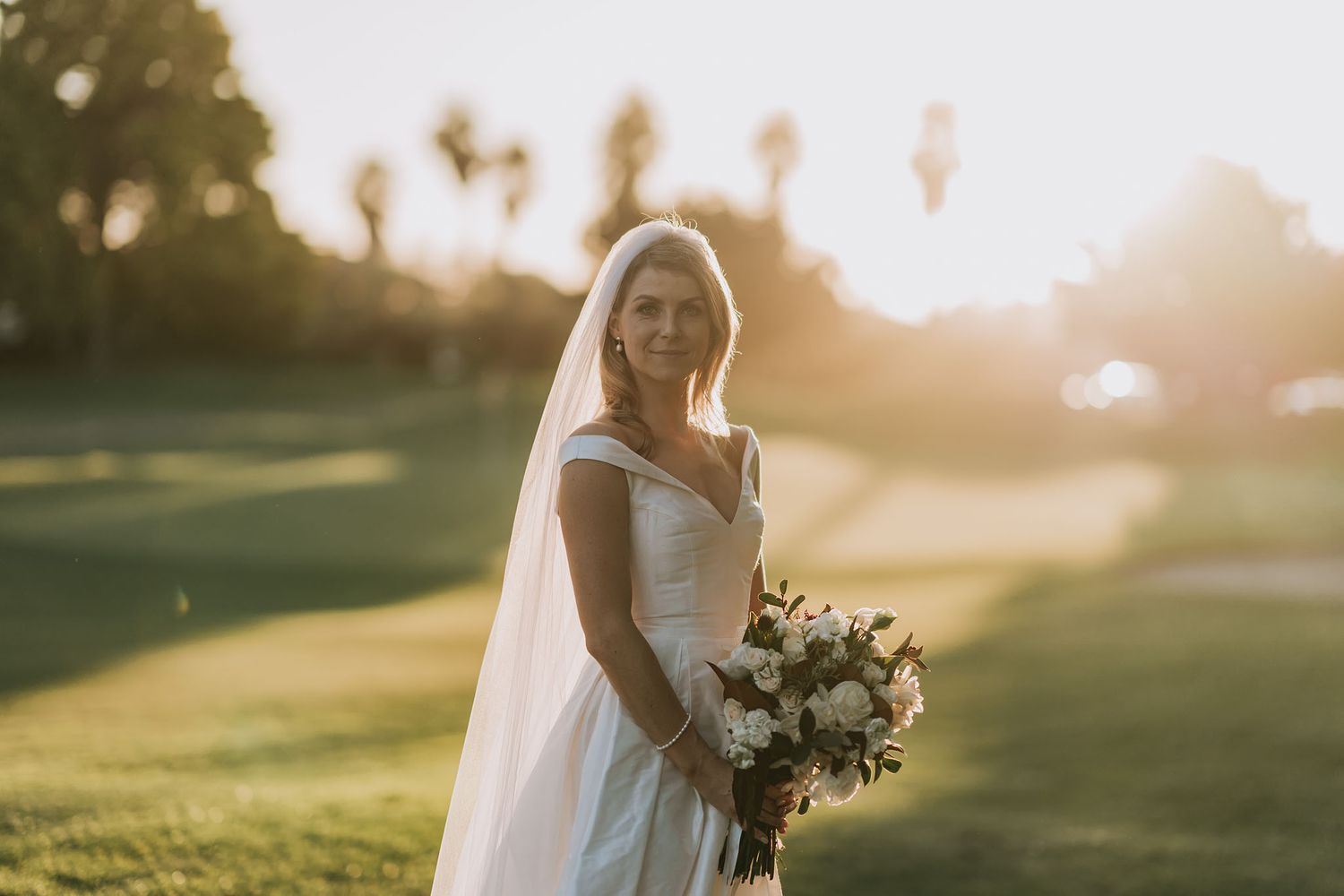 A bride in a classic white wedding dress holds a bouquet while standing in a sunlit field at golden hour.