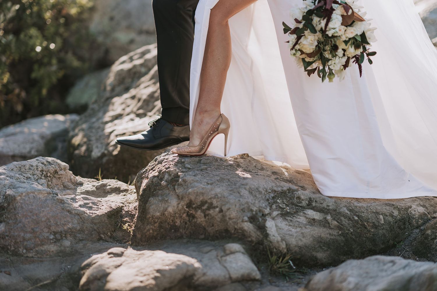 Close-up view of bride wearing elegant heels standing on rocks with white wedding dress and bouquet visible.