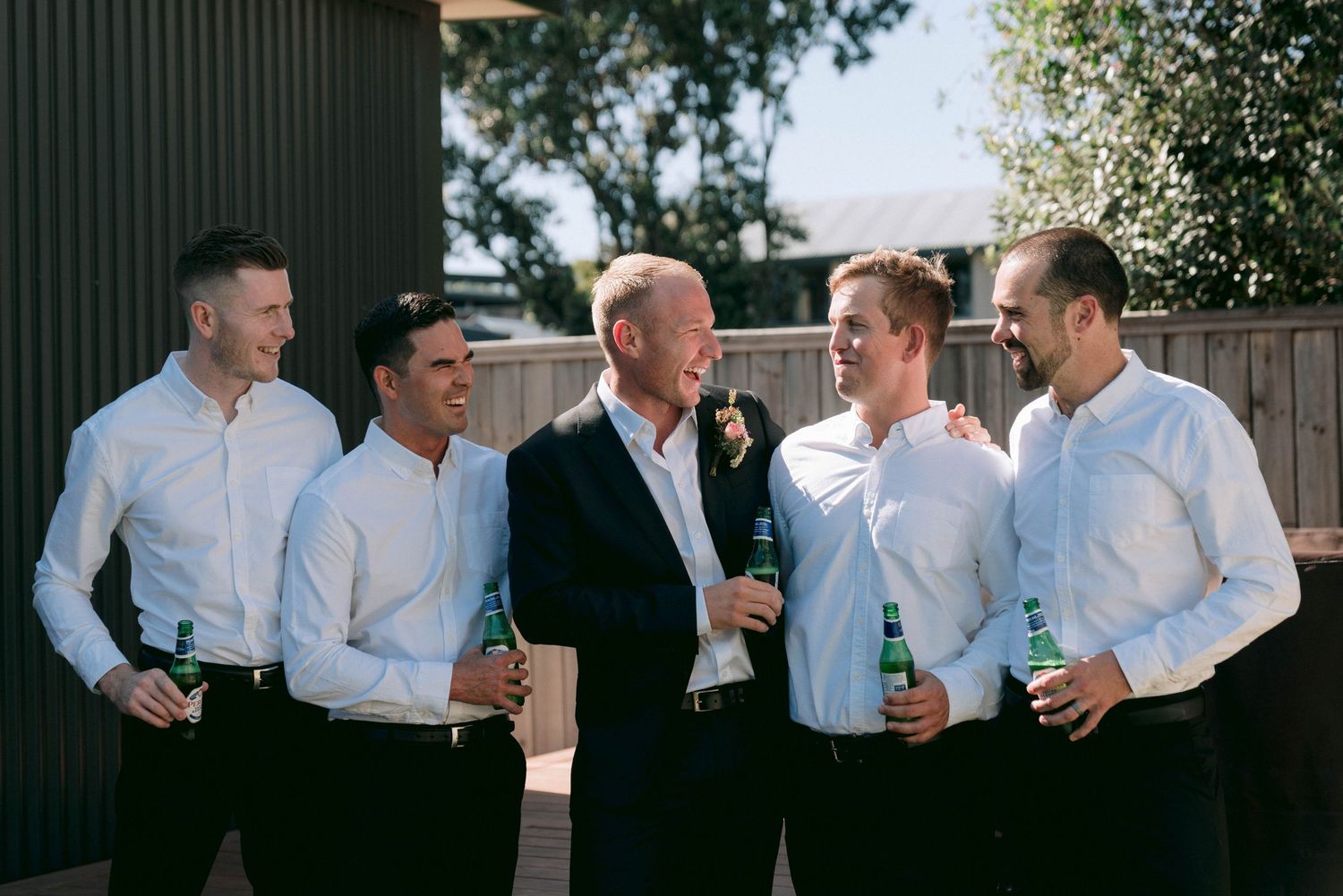 A group of men in white shirts share drinks and laughter outdoors during a social gathering.