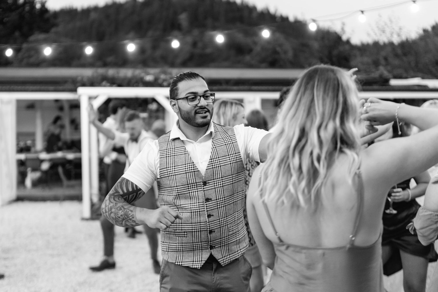 A guest in a waistcoat stands at an outdoor event venue with string lights visible in the background.