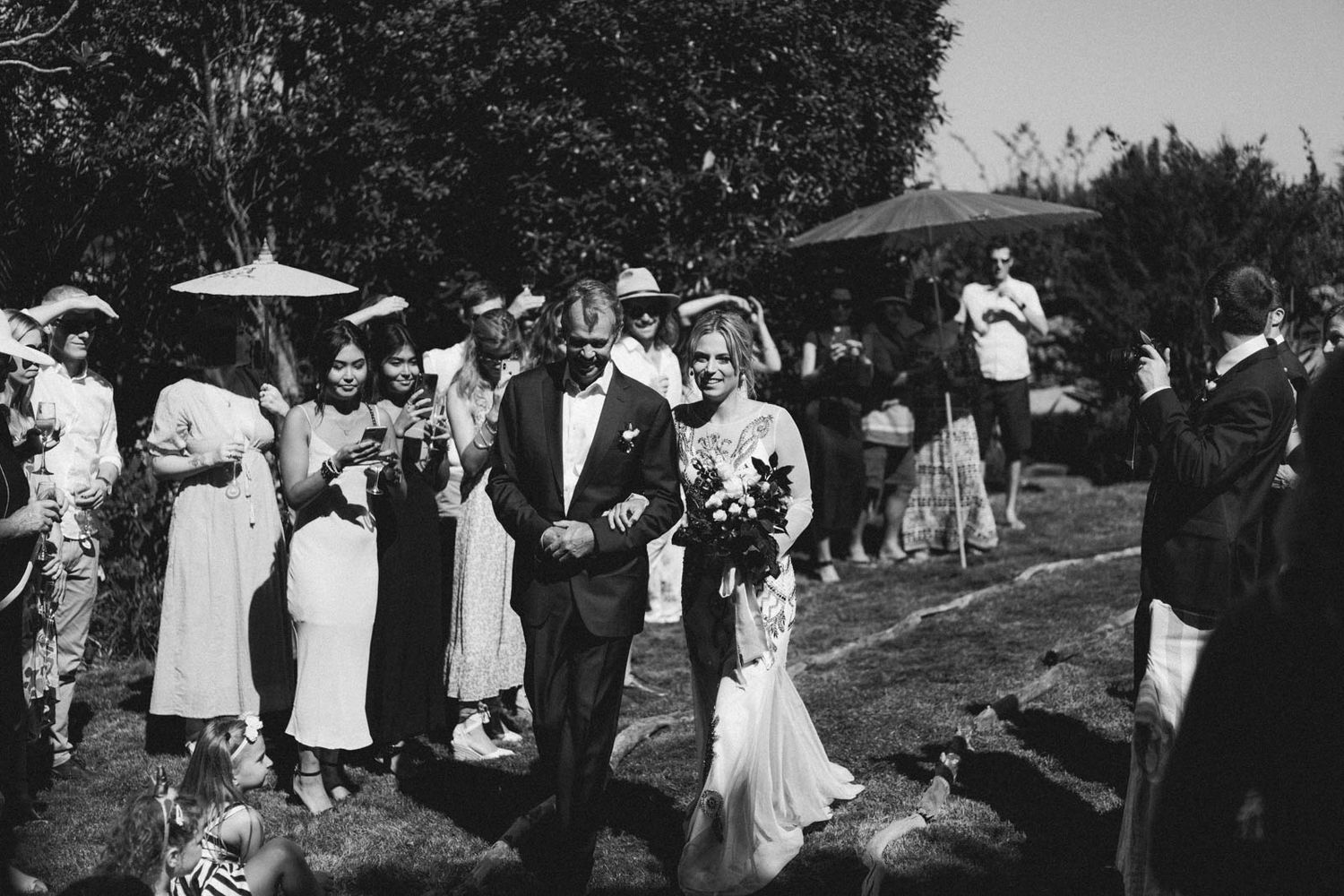 A group of wedding guests and partygoers in white attire gather outdoors under a shaded canopy during a celebration.