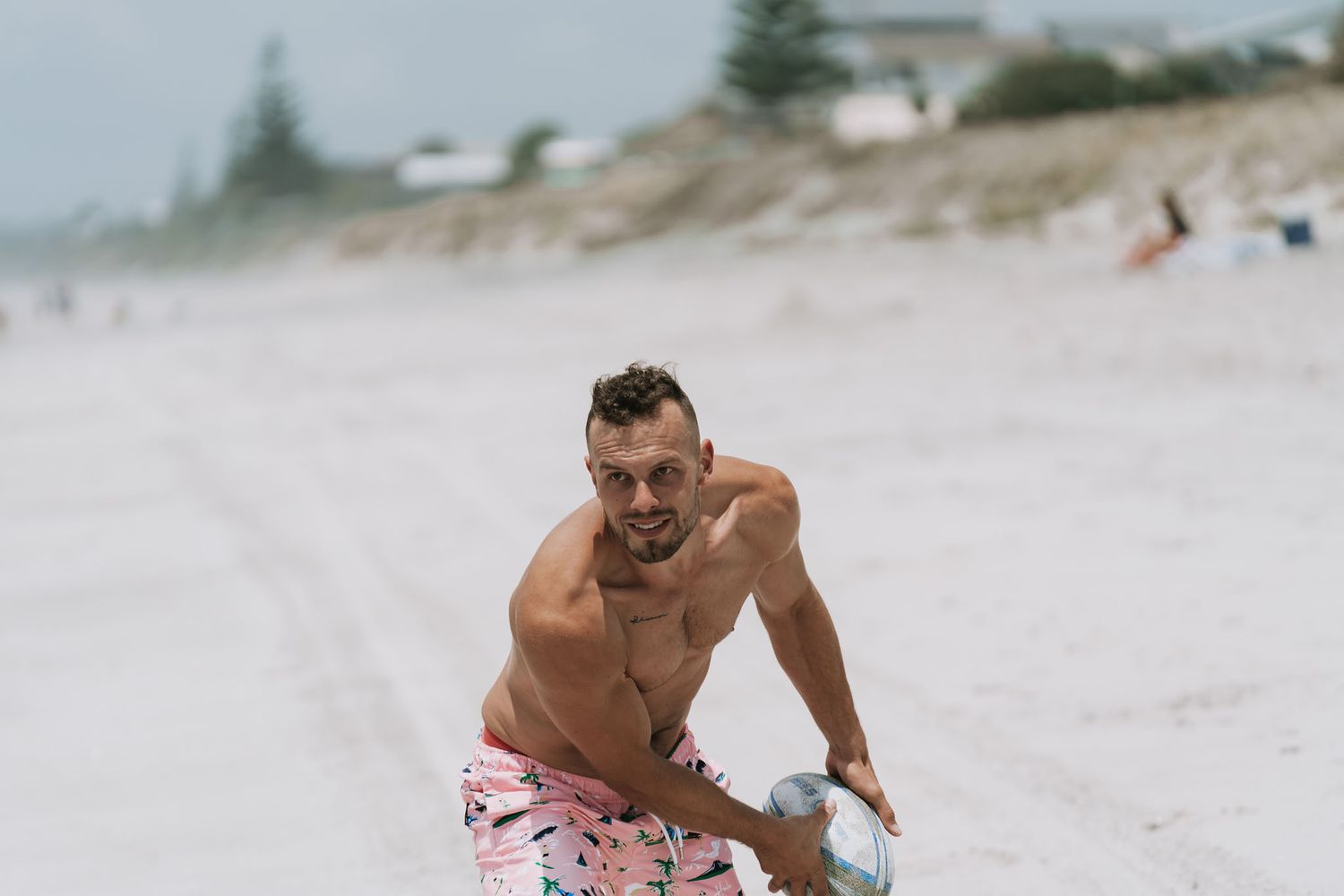 A shirtless person in floral swim trunks kneels on a sandy beach with blurred coastal background.