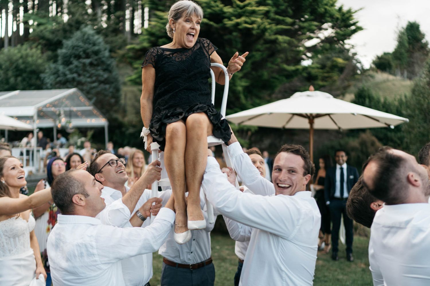 Wedding guests participate in a synchronized dance line on an outdoor dance floor under trees.