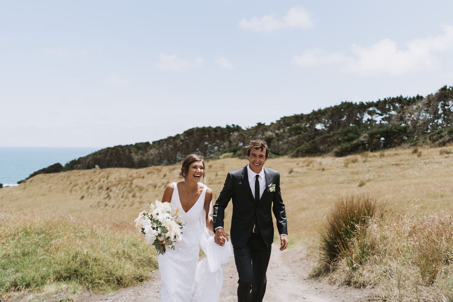 Newlyweds hold hands and walk together through a scenic coastal grassland with mountains in the background.