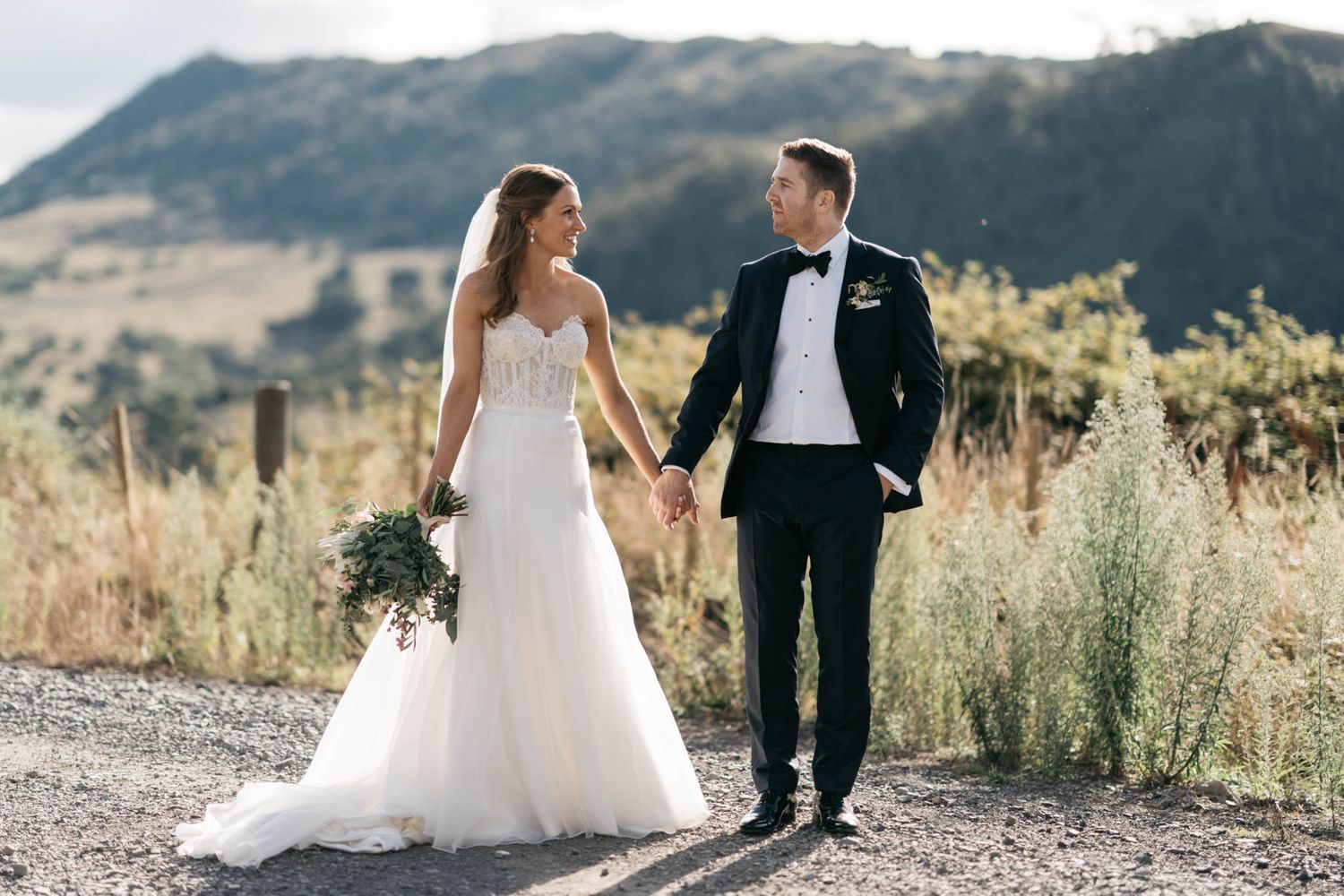 Newlyweds holding hands on a mountain path with scenic rolling hills and golden sunlight in the background.