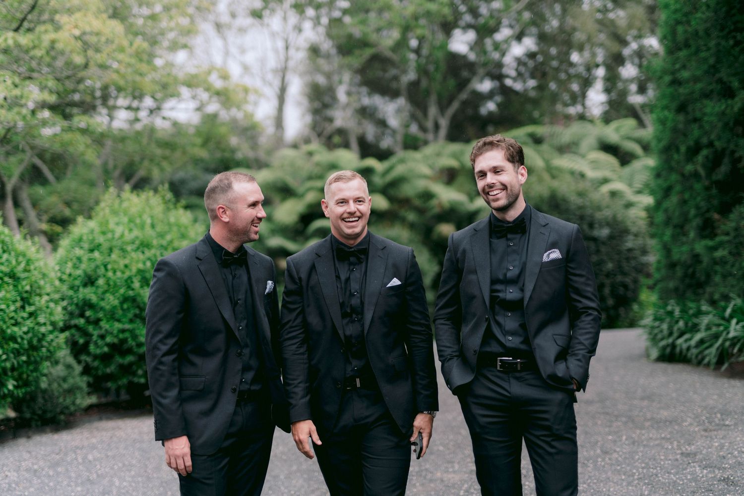Three men in black suits share a candid moment on a garden path.
