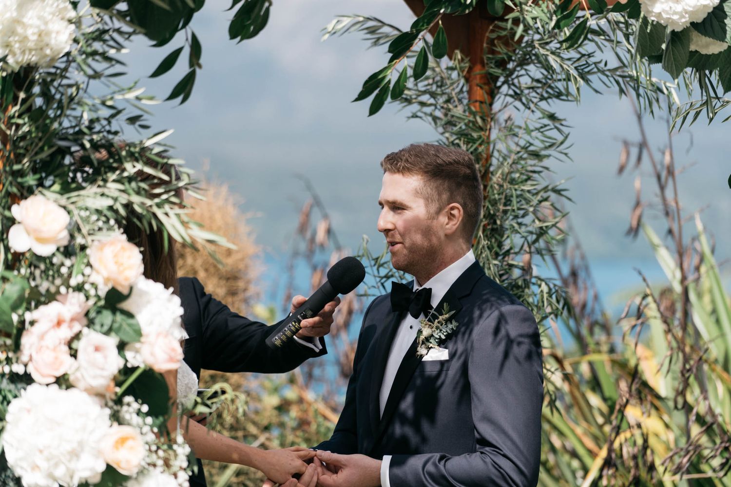 Someone in a black suit speaks at a microphone under an elegant floral arch with white flowers and greenery.