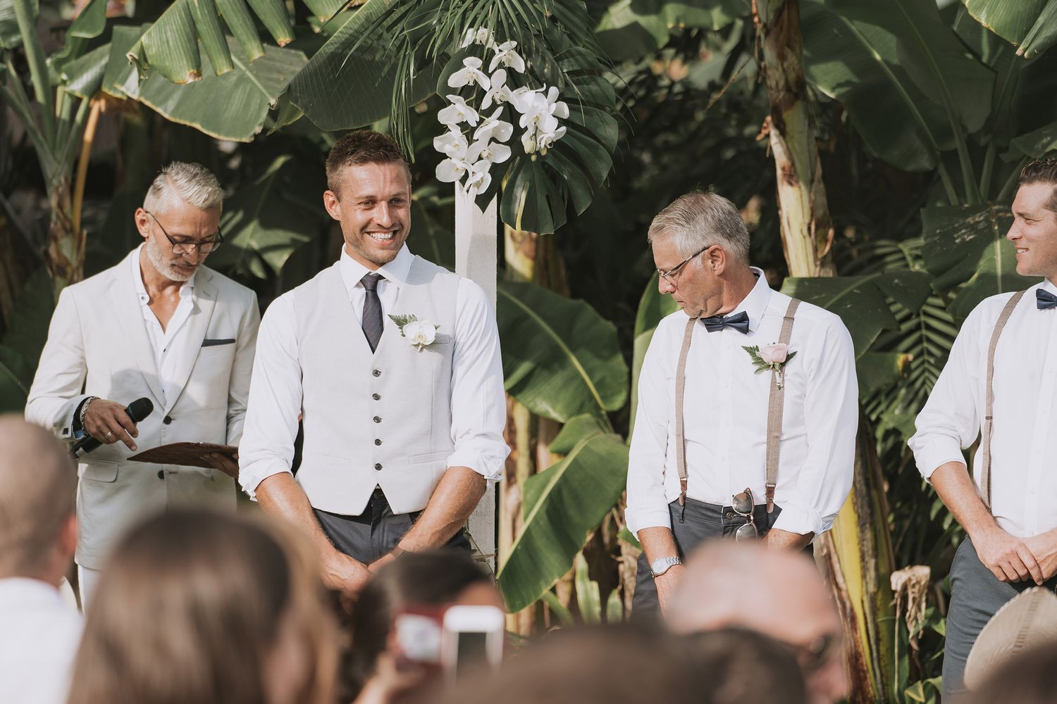 Wedding ceremony in progress under tropical palm trees with attendees in white attire.