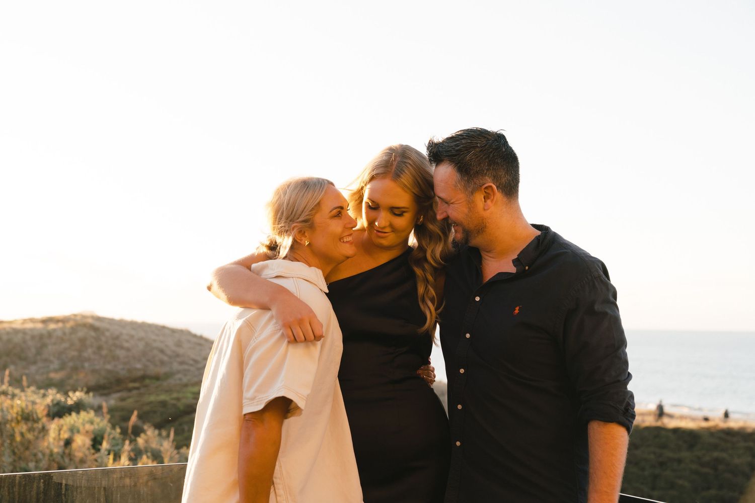 Three people hugging and laughing together at sunset with coastal mountains in the background.