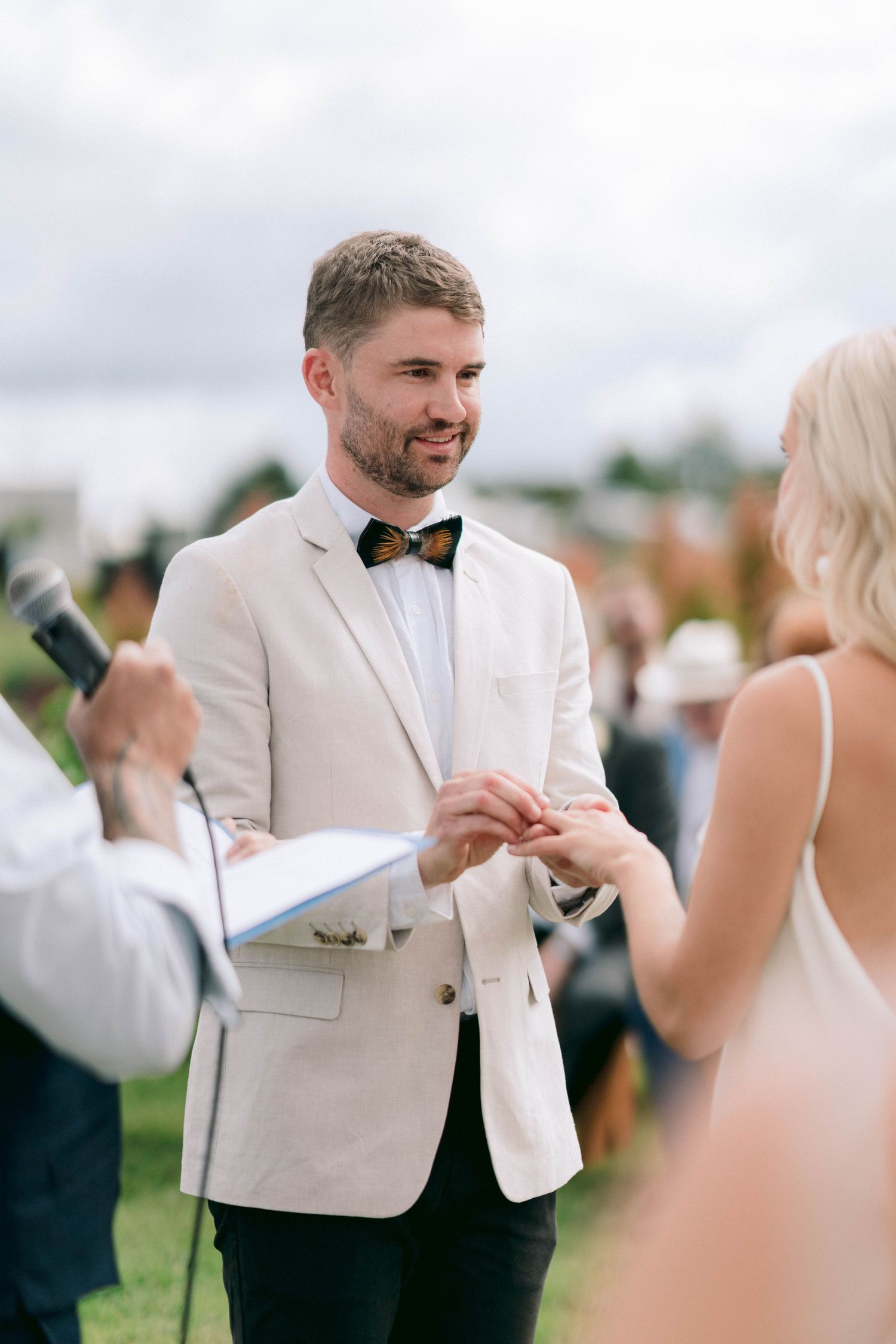 Exchange of rings during outdoor wedding ceremony with white blazer and bow tie.