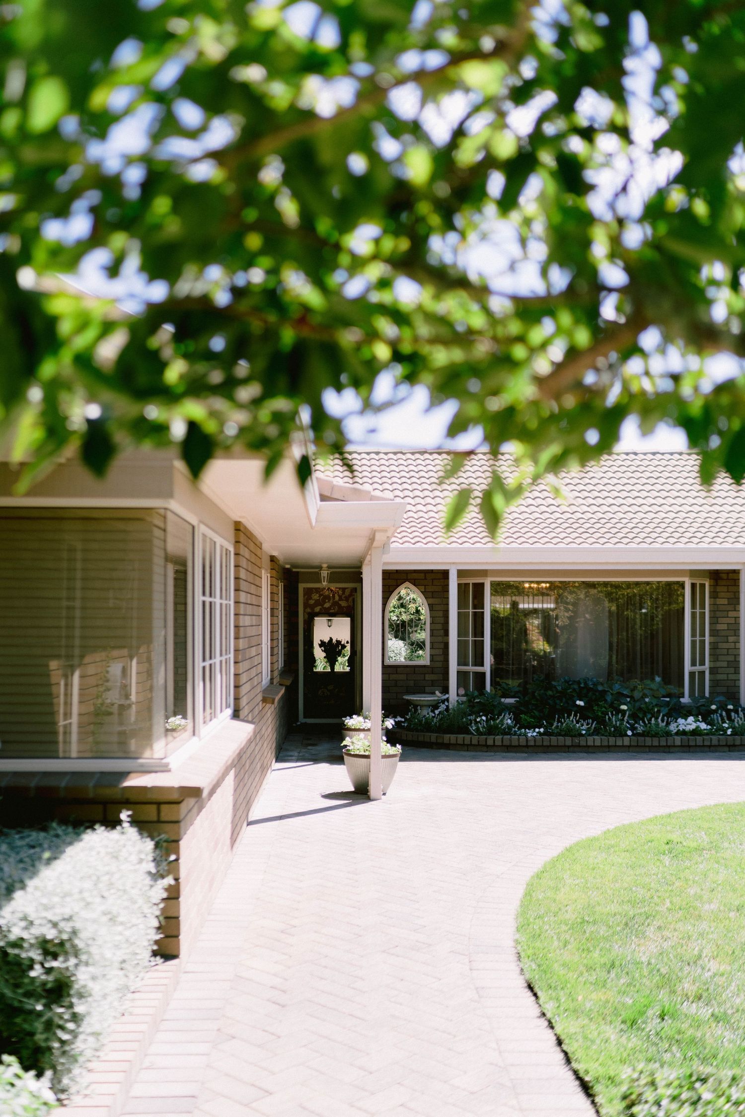 Residential house exterior with concrete walkways and green landscaping under shaded tree canopy in suburban setting.