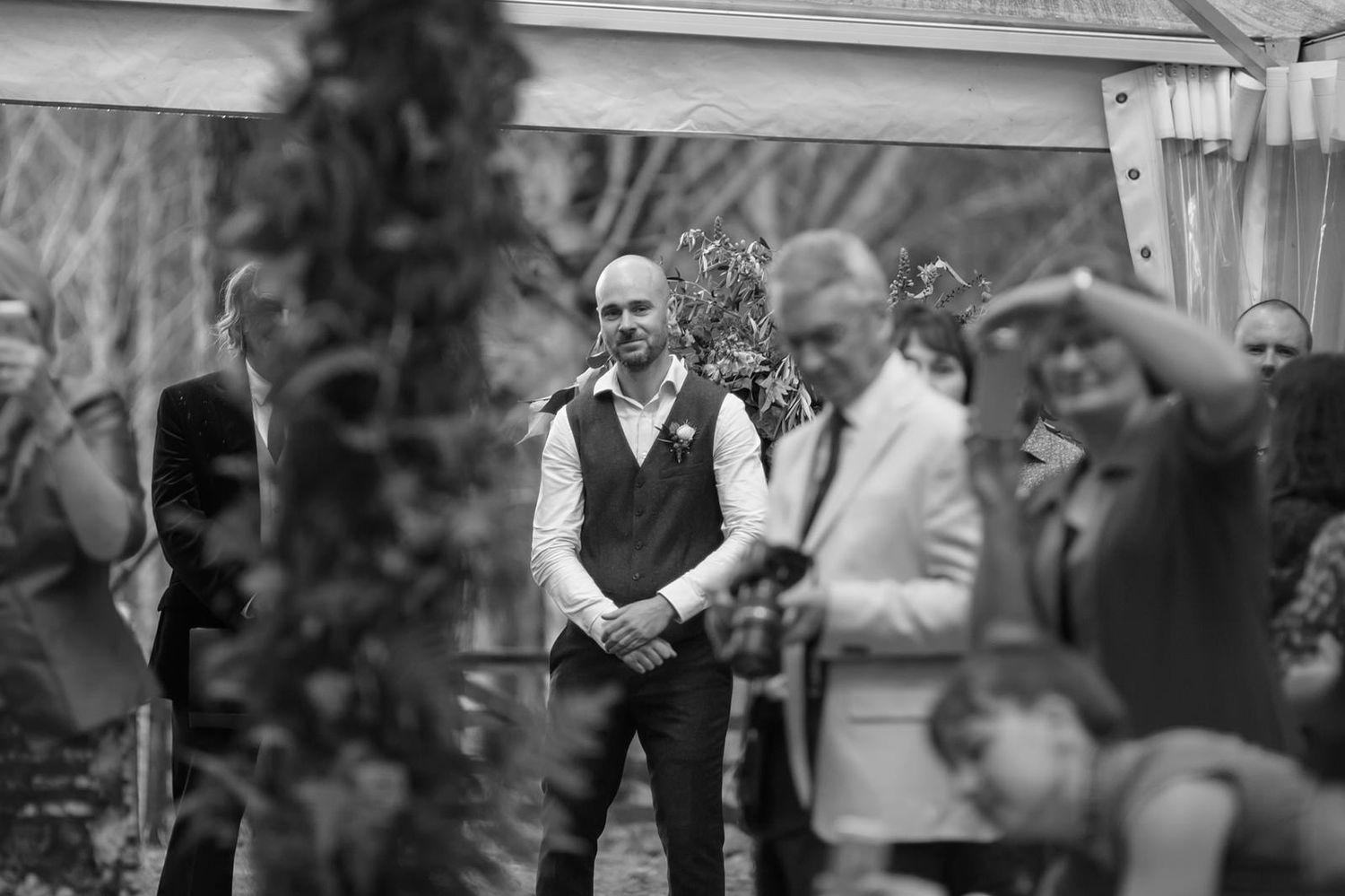 A black and white candid photograph captured during an outdoor wedding ceremony under a tent.
