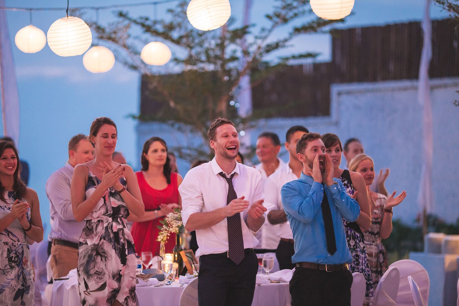 Evening outdoor reception with guests clapping under hanging paper lanterns.