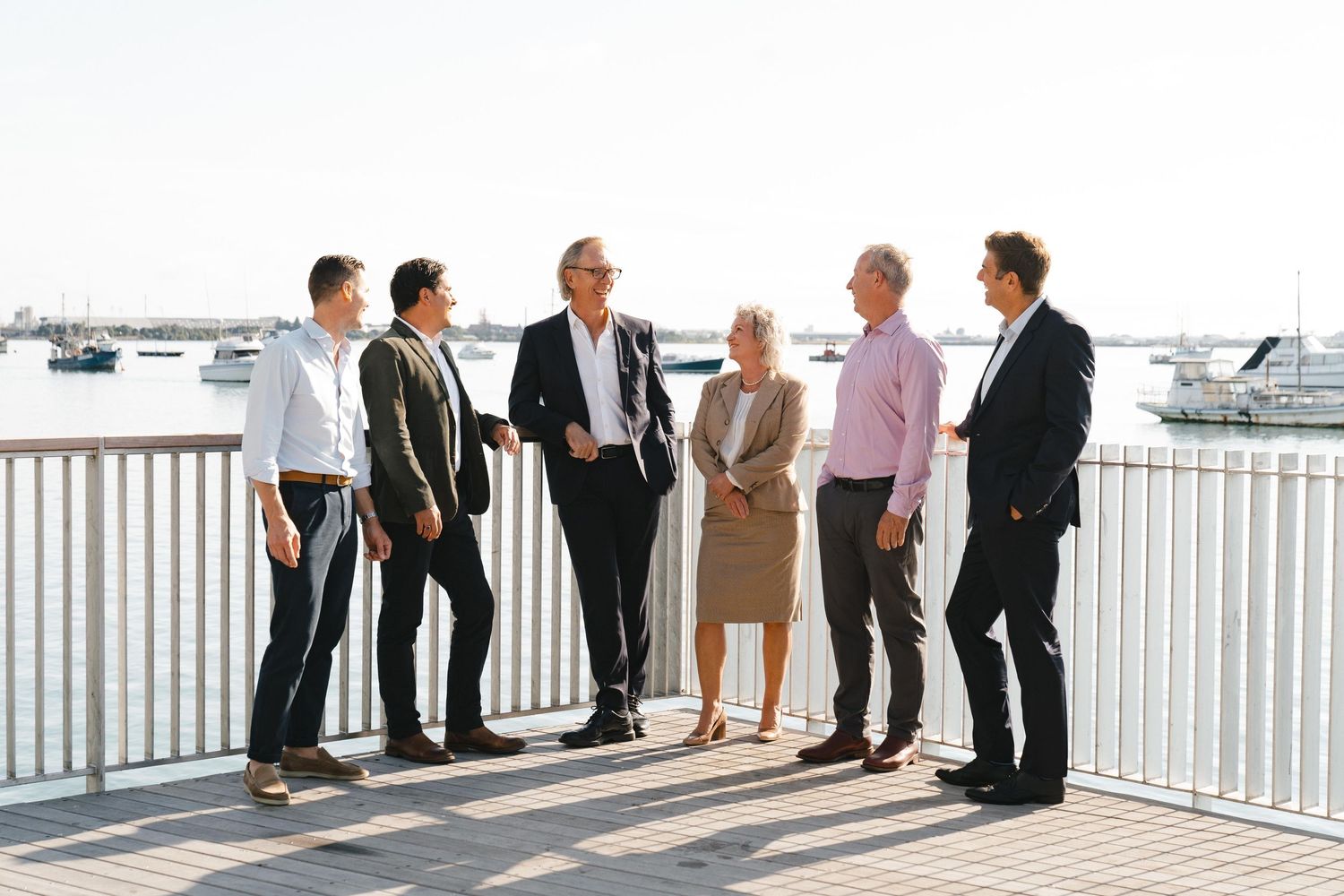 People gathered on an outdoor deck with white railings and boats visible in the background at a waterfront venue.
