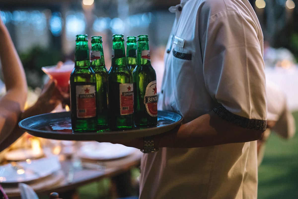 Server carries a tray of green glass beer bottles at an outdoor event.