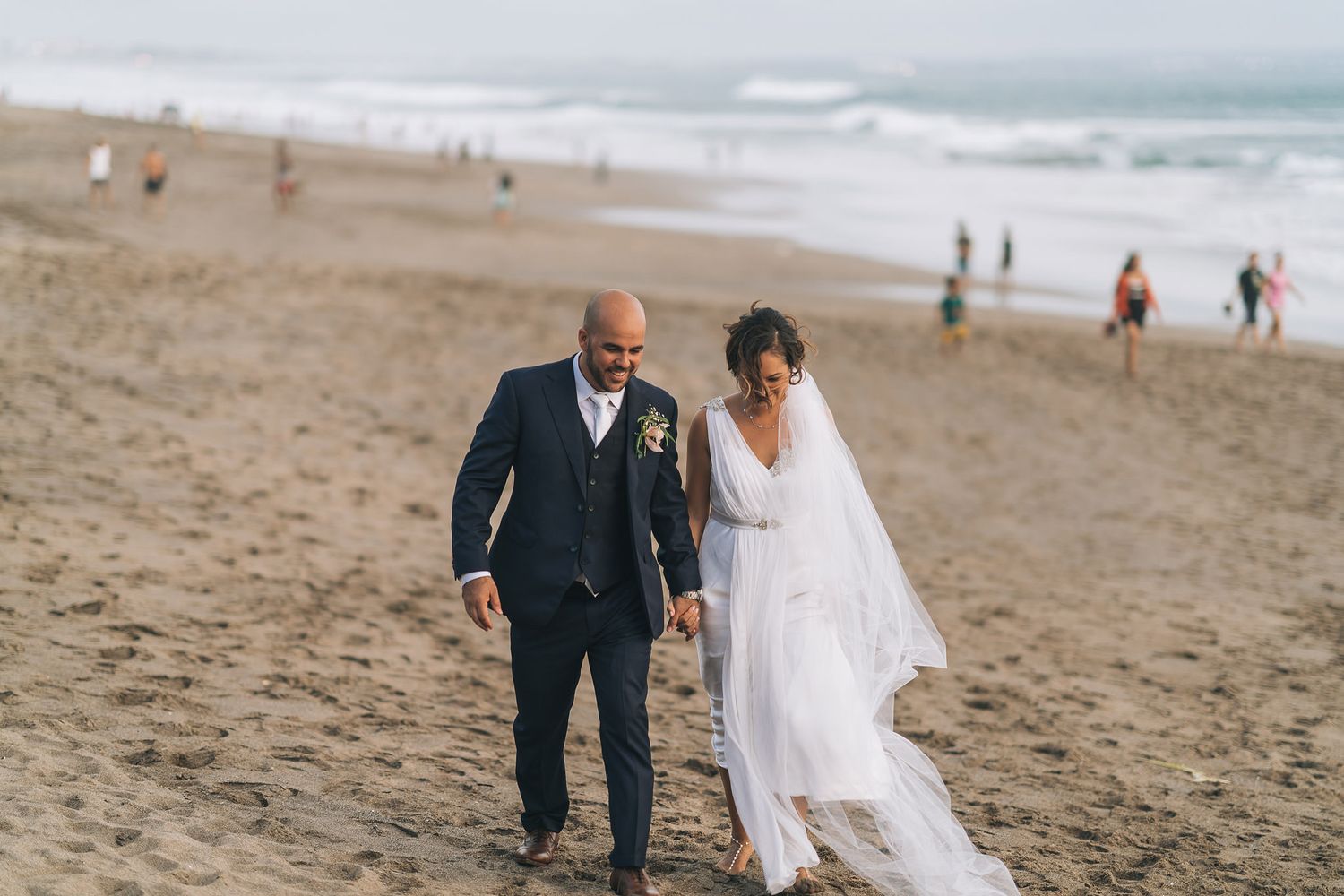 Couple walking hand in hand along a scenic beach shoreline in formal wedding attire.