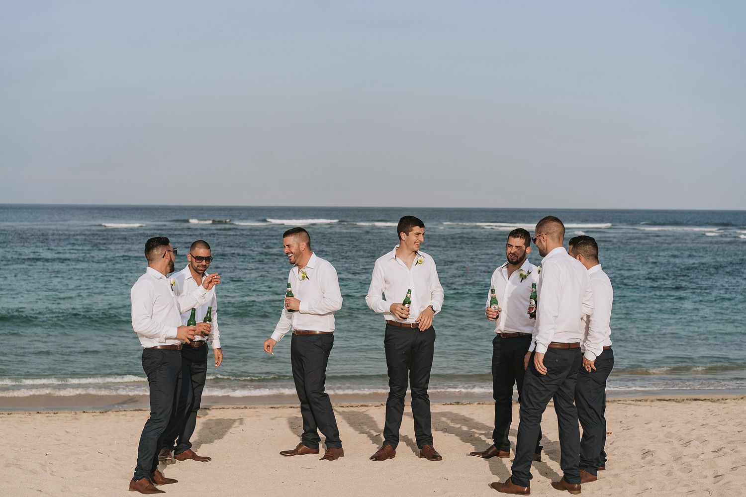 Groomsmen in white shirts gather on a beach with ocean waves in the background.