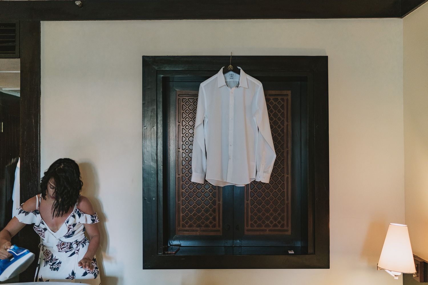 A white dress shirt hangs framed on a dark wall next to a table lamp in an interior setting.