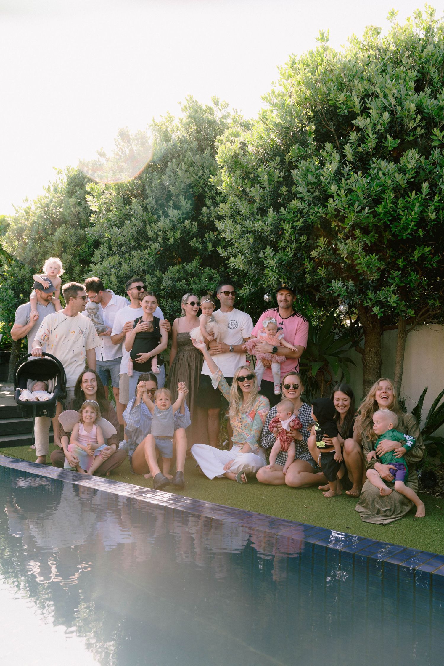 Large group of friends sitting along the edge of a swimming pool at an outdoor party.