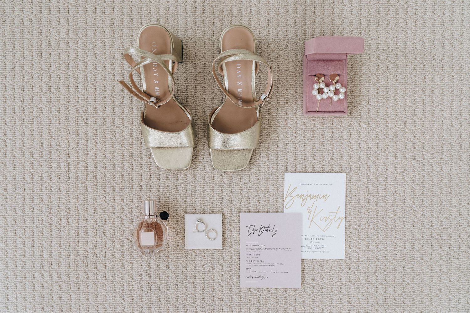 White and gold wedding accessories including rings, earrings, and shoes arranged on a textured neutral background.