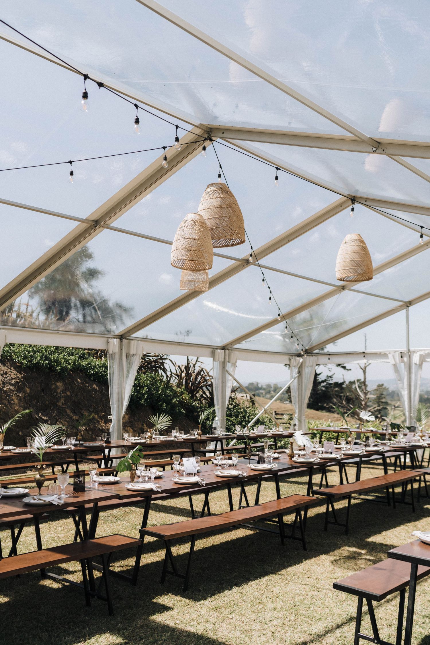 Wedding reception setup under clear tent with woven pendant lights and long wooden tables on grass.