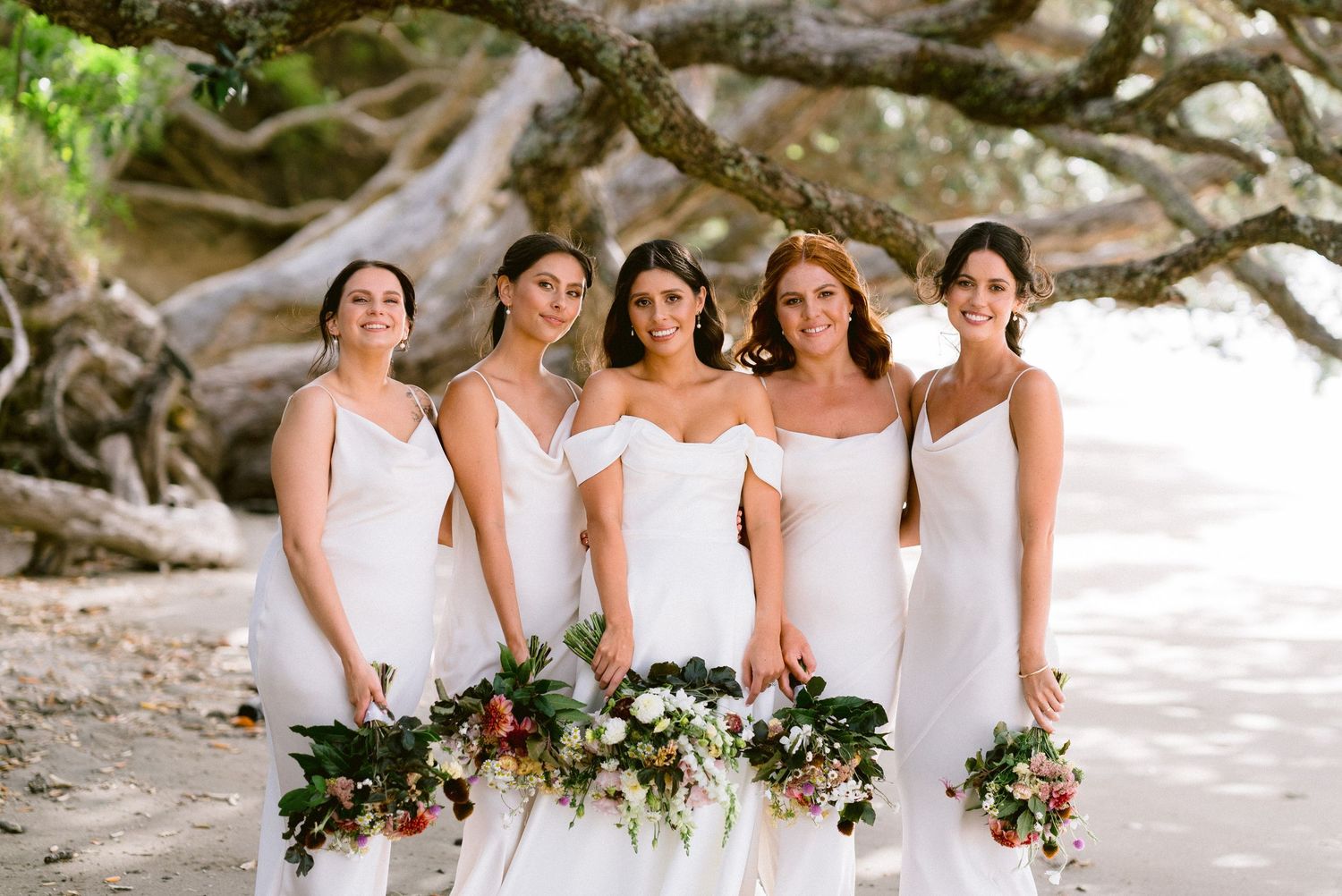 Bridesmaids in white dresses holding bouquets pose together on beach with tree backdrop.